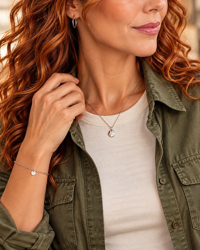 Detailaufnahme einer feinen Silberkette mit rundem Anhänger, einer silbernen Creole und eines Armbands zu Aimonis olivgrüner Bluse.